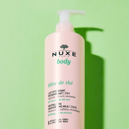 En rosa flaske med Nuxe body lotion med pumpe, grønn og svart tekst, og en skygge på en grønn bakgrunn.