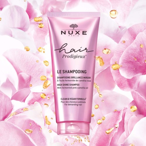 Pinkfarbige Shampoflasche mit Gold und Blütenblättern im Hintergrund, beschriftet mit 'Nuxe Hair Prodigieux' und 'High Shine Shampoo'.
