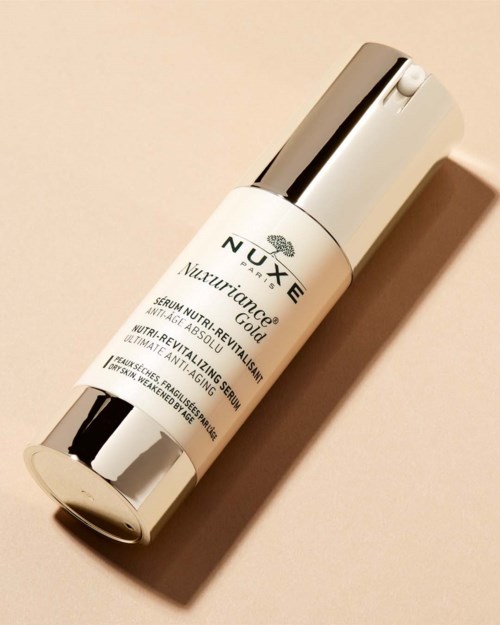 En hvid flaske med sølvkapsel, mærket 'NUXE Nuxuriance Gold,' indeholdende anti-aging serum. Den placeres på en lys baggrund.