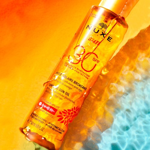 En guldfärgad flaska av Nuxe Sun SPF 30 sololja med blommig design, placerad på en gul och blå bakgrund.
