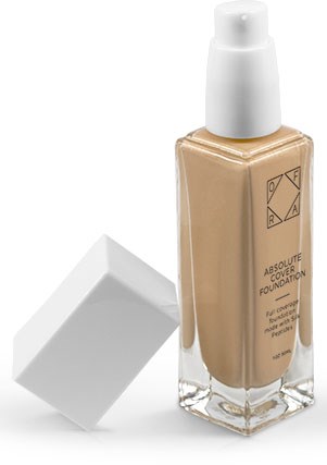 En beige vätskefondationsflaska med vit pump och vit kork, ligger bredvid en vit fyrkantig låda.