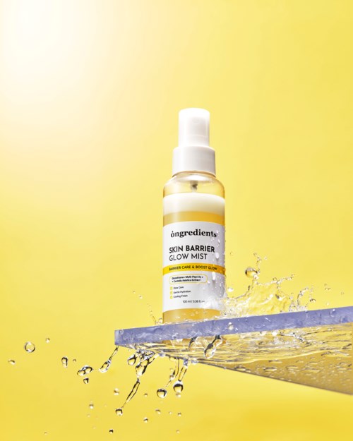 Butelka sprayu oznaczona 'Skin Barrier Glow Mist' na pływającej platformie z rozpryskami wody, żółte tło.
