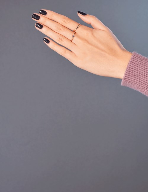 En hand med svarta lackade naglar och en ring, bär en rosa ärm, sträcker sig mot en grå bakgrund.