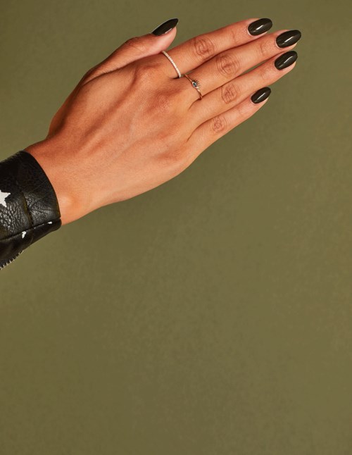 Hand with dark nails and rings, wearing a black wristband, against a plain background.