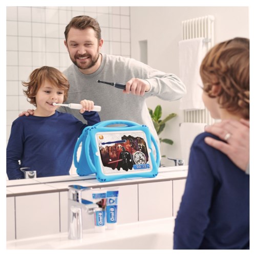Vader helpt zoon tanden te poetsen in de badkamer, met een tablet die een tekenfilm toont op de wastafel.
