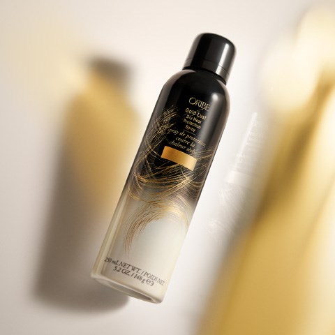 En svart og gullflaske med Oribe tørr teksturiserende spray med et silkeaktig virveldesign. Skygger og lys fremhever flasken.