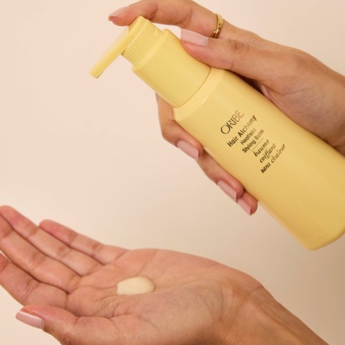 En person bruker en gul flaske med Oribe Heat Styling Spray, holder den over sin åpne hånd med en klatt av produktet.