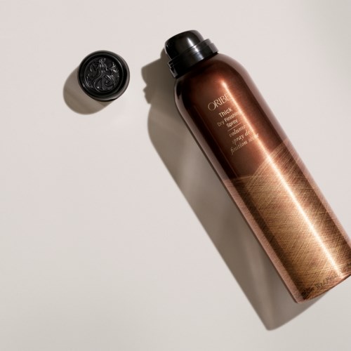 A brown bottle of Oribe hair spray lying on a white surface with a black cap nearby.