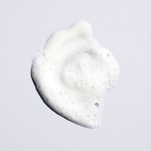 A blob of white foam or soap with bubbles, looks like a splash, on a plain gray background.