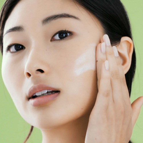 En kvinna applicerar kräm eller lotion på sitt ansikte med sin hand, mot en grön bakgrund.