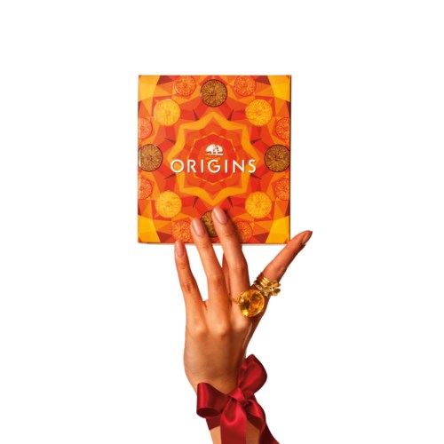 A hand with rings and a red ribbon holds a colorful, geometric-patterned box labeled 'ORIGINS'.