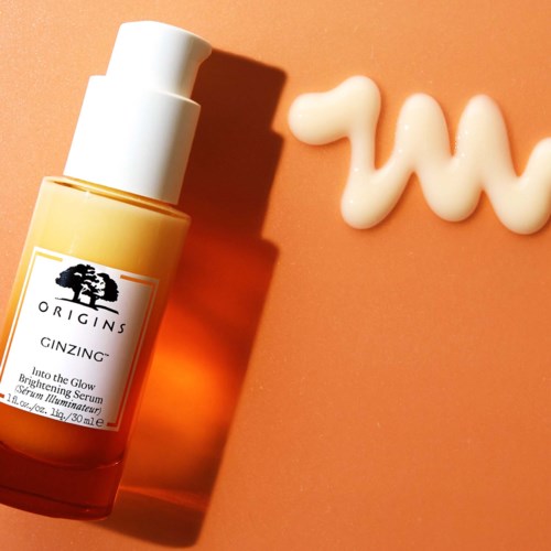 En gul flaska med Origins GinZing serum med vit kräm eller lotion bredvid på en rosa bakgrund.
