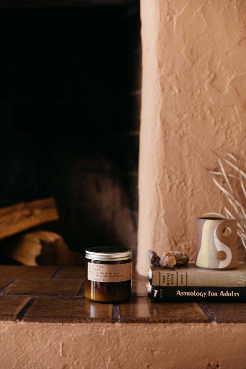 Een potje, een boek getiteld "Astrology for Adults," en een vaas met gedroogde planten staan op een houten oppervlak naast een gestructureerde beige muur.