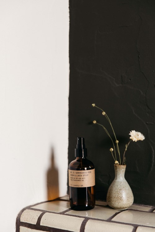 Eine braune Sprühflasche und eine kleine Vase mit weißen Blumen stehen auf einer gefliesten Oberfläche vor einer schwarz-weißen Wand.