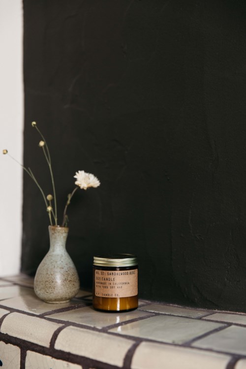 Eine kleine Vase mit weißen Blumen neben einem Glas auf einer ziegelbedeckten Oberfläche vor einer schwarzen Wand.