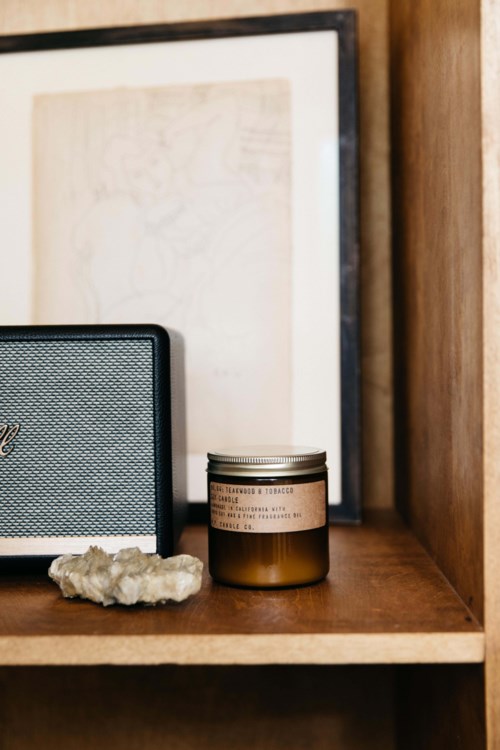 A small speaker, a candle, and a rock sit on a wooden shelf with framed artwork behind them.