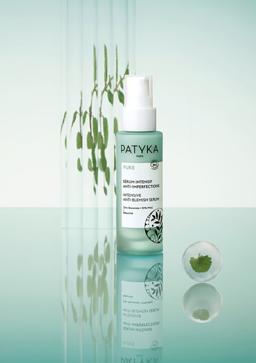 En flaske af Patyka Pure Anti-Imperfection Serum med et klart, minimalistisk design og hvid pumpe, placeret på en reflekterende overflade.