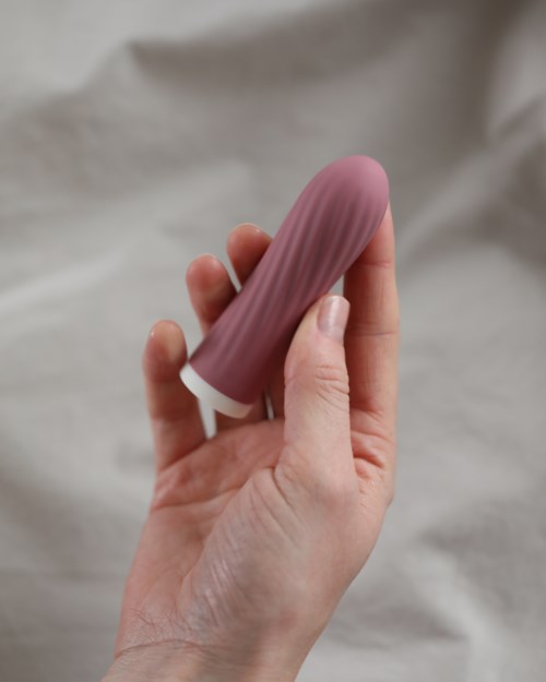 Henkilö pitää kiinni violettia, peniskuvioista esinettä, sormet tarttuvat siihen. Tausta on vaalea ja yksinkertainen.