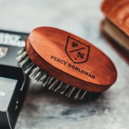 Eine Holzrasierbürste mit Percy Nobleman-Logo und Metallborsten auf einer Oberfläche.