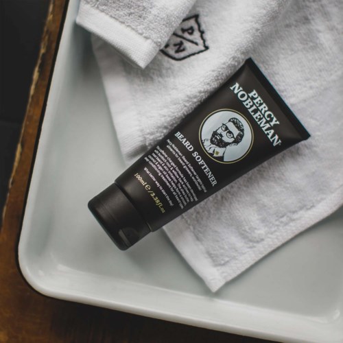 Eine schwarze Tube mit der Aufschrift 'Beard Softener' liegt auf einem weißen Handtuch auf einem Tablett.