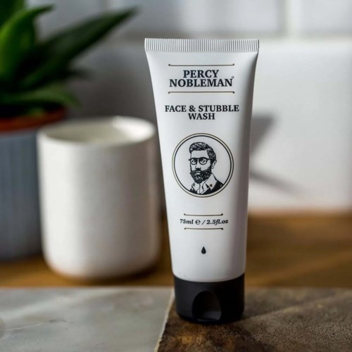 En vit tub med ansiktsrengöring med svart lock, märkt Percy Nobleman, på en träyta, bredvid en grön växt.
