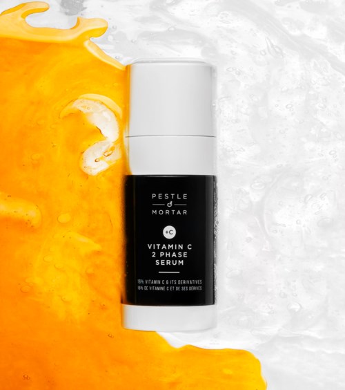 Czarna i biała butelka oznaczona 'Vitamin C 2 Phase Serum' leży na powierzchni z pomarańczową i białą cieczą.