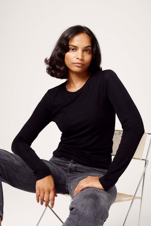 A woman wearing a black sweater and gray jeans sitting on a white chair against a plain light background.