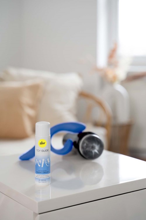 Eine blaue und weiße Flasche mit Pjur Gleitmittel auf einem weißen Nachttisch, im Hintergrund eine blaue Massagegerät und Bett.