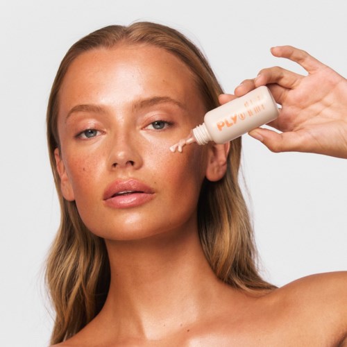 En kvinna med långt hår applicerar lotion på ansiktet. Hon har ett neutralt uttryck och tittar på kameran. Bakgrunden är vit.