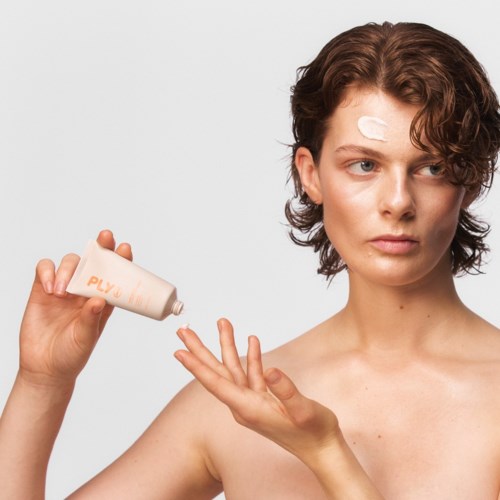 En kvinna smörjer in sin hand med lotion, med ett plåster på pannan, mot en enkel bakgrund.