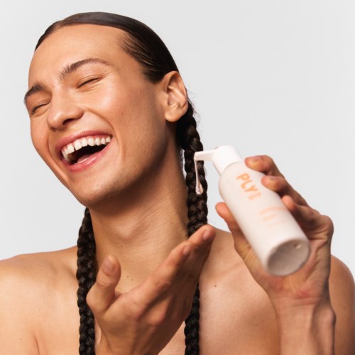 En leende person med långa flätor använder lotion på ansiktet med en vit flaska.