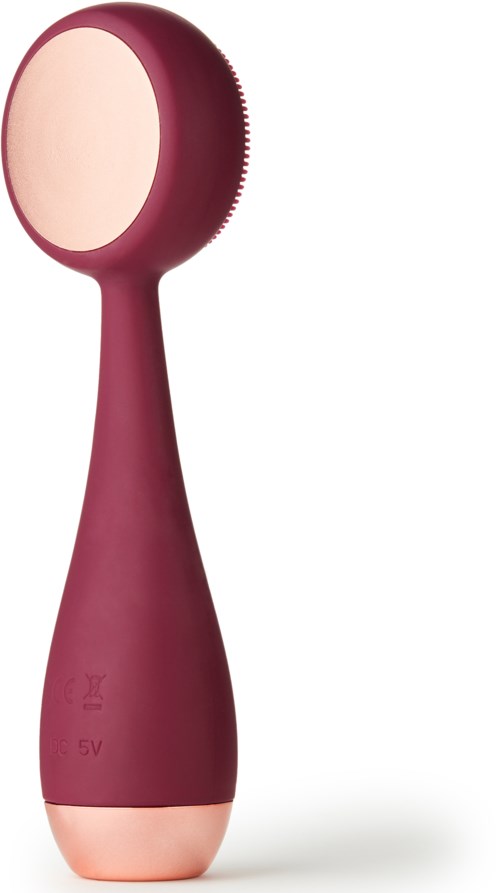 A handheld facial cleansing device with a large round brush head and purple handle.