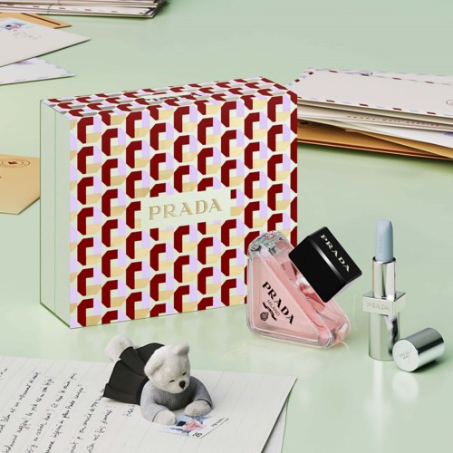 Prada parfume flaske med pink væske og sort låg, ved siden af en læbestift og en lille bamse på et skrivebord.