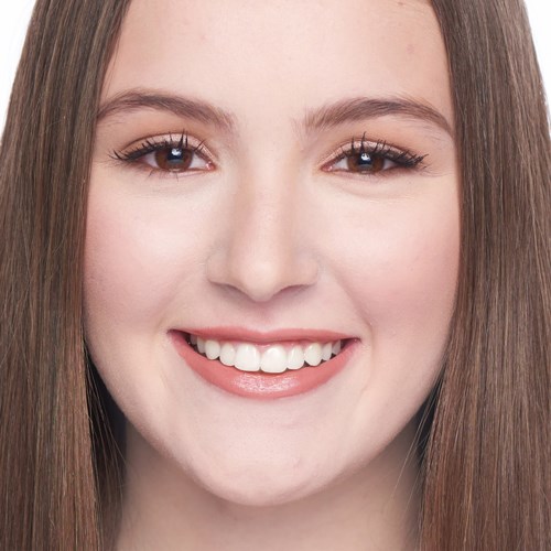 A young woman with long brown hair smiling, showing her teeth, and clear skin.