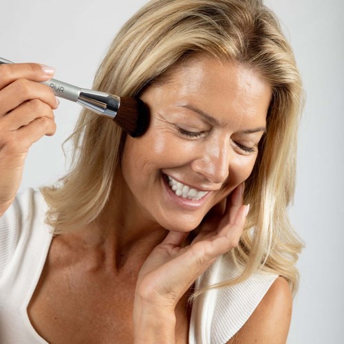 Eine lächelnde Frau mit geschlossenen Augen, die Makeup mit einem Pinsel auf die Wange aufträgt.