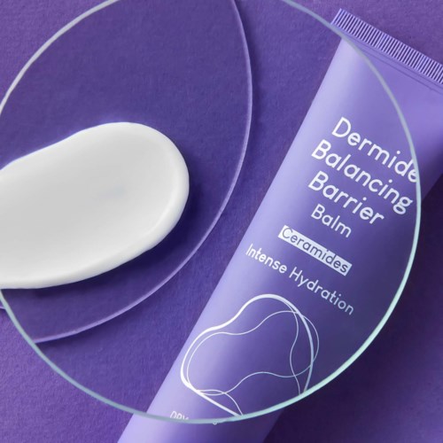 En lila tub med Dermide Balancing Barrier Balm med etikett, placerad på ett transparent glasfat, med en vit substans bredvid.