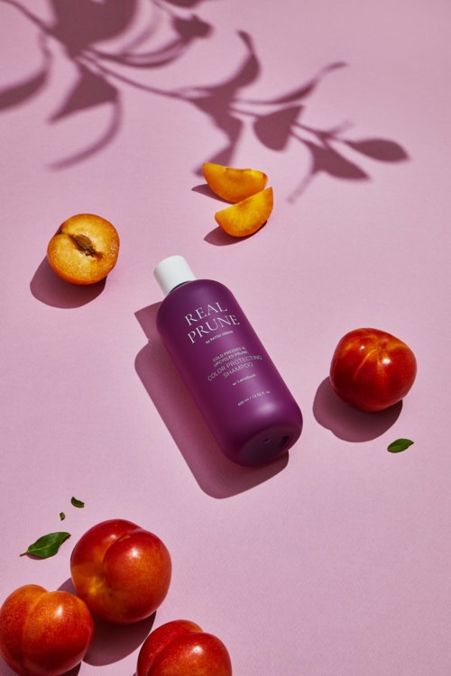 Eine lilafarbene Flasche mit 'Real Prune' auf, umgeben von Pfirsichen, Blättern und orangefarbenen Kernen auf pinkem Hintergrund.
