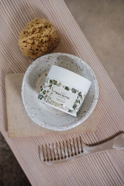A jar of body butter on a small plate, a sponge, and a fork on a pink striped cloth.