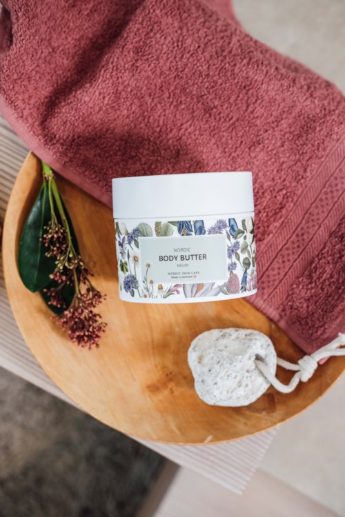 A body butter jar on a wooden tray with a red towel, a small green plant, and a white pumice stone.