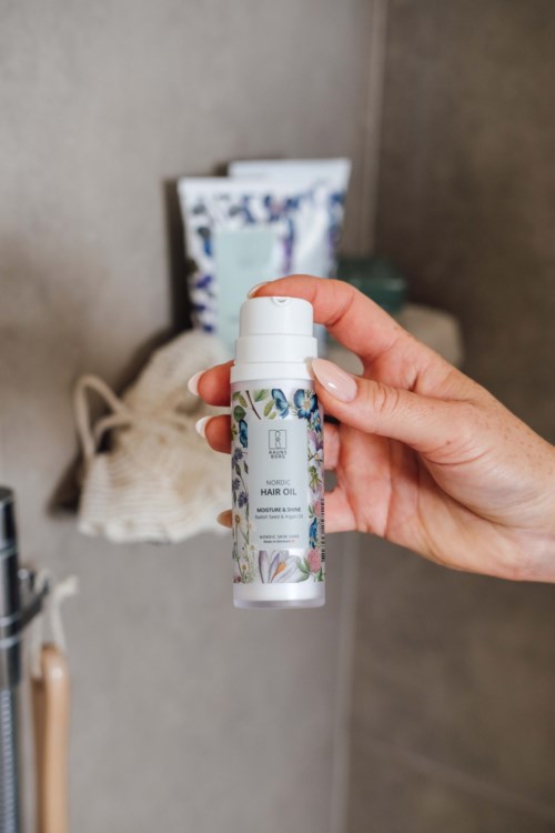 Dłoń trzyma małą białą butelkę z napisem 'HAIR OIL' w pomieszczeniu z półkami i przedmiotami w tle.