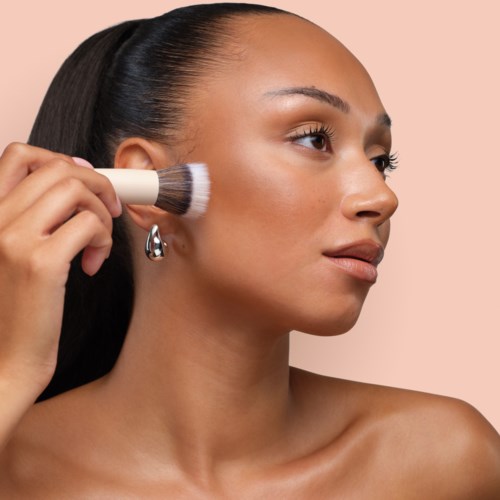 Een vrouw brengt make-up aan met een kwast op haar gezicht, kijkt opzij, met een heldere huid en lang donker haar.