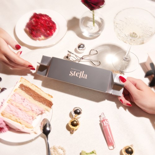 Ett presentförpackning med etiketten 'Stella' hålls av händer med röda naglar. Det finns tårta, en drink och dekorativa föremål på bordet.