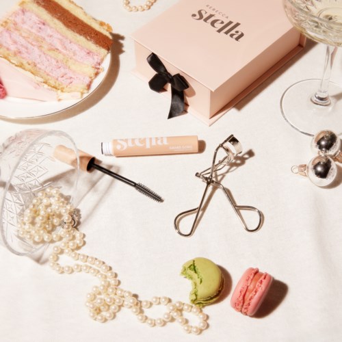 En platt bild med skönhets- och festartiklar, inklusive tårta, smycken, smink, macarons och ett glas.