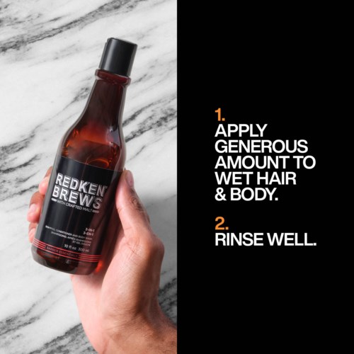 En hånd holder en brun flaske med Redken Brews hårpleieprodukt på en hvit marmorflate, med bruksanvisning på svart bakgrunn.