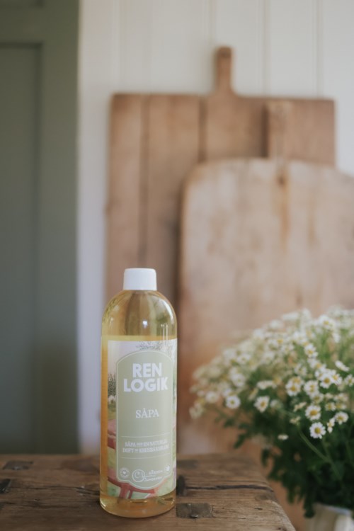 Een fles schoonmaakmiddel met het label 'REN LOGIK SÄPA' op een houten tafel, met bloemen in een potje in de buurt en een houten bord op de achtergrond.