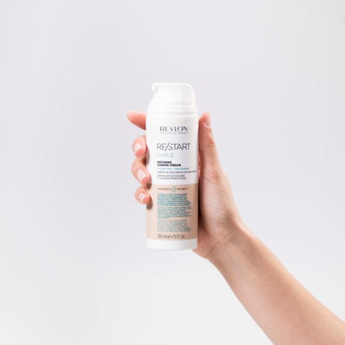 A hand holds a white bottle of Revlon hair product against a plain background.