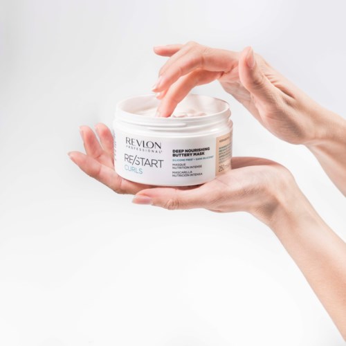 A person uses fingers to scoop out cream from a white jar labeled Revlon hair mask.