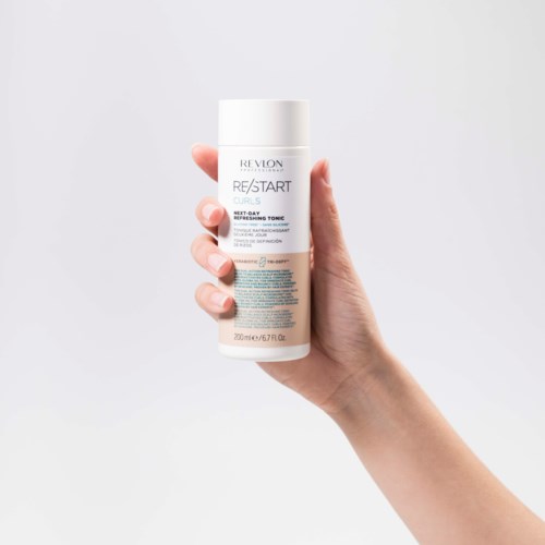 En hand håller en vit flaska med blå text för Revlon Re/Start hårtonerprodukt. Enkel bakgrund.