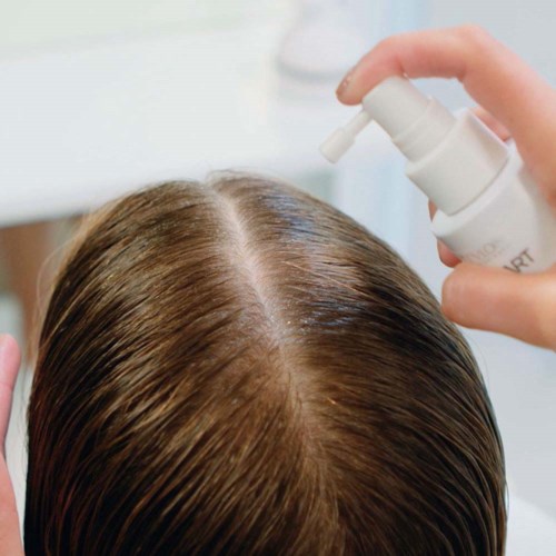 A person sprinkles hair product on their scalp, focusing on the part of brown hair.