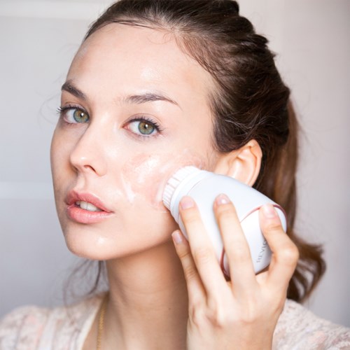 A woman uses a facial cleansing brush on her cheek, with some foam visible. She has light skin and brown hair.
